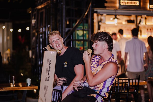 Two people sitting at a table with a chair labeled "Outdoor Foldable Chair" and a can of beer on the table.