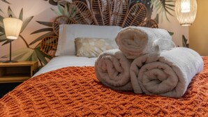 Ein ordentlich bezogenes Bett mit aufgerollten weißen Handtüchern, einer strukturierten orangefarbenen Decke und einer Wand mit tropischem Blattmuster.