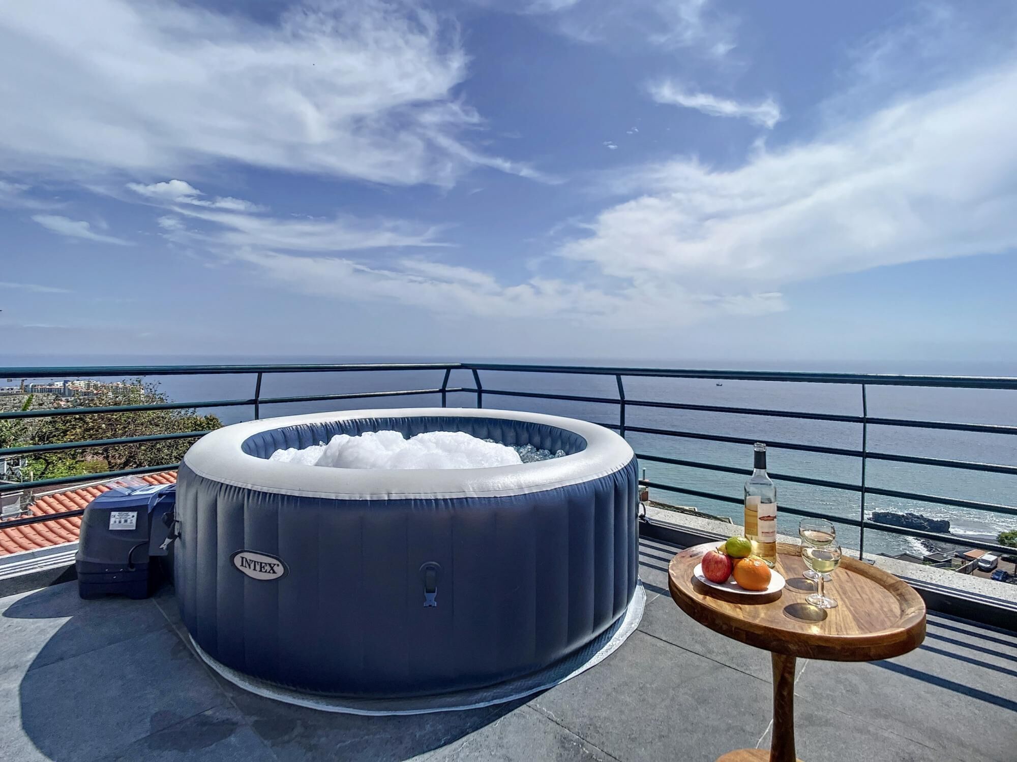Een opblaasbaar bubbelbad op een balkon met uitzicht op zee, een fles wijn en een schaal met fruit.