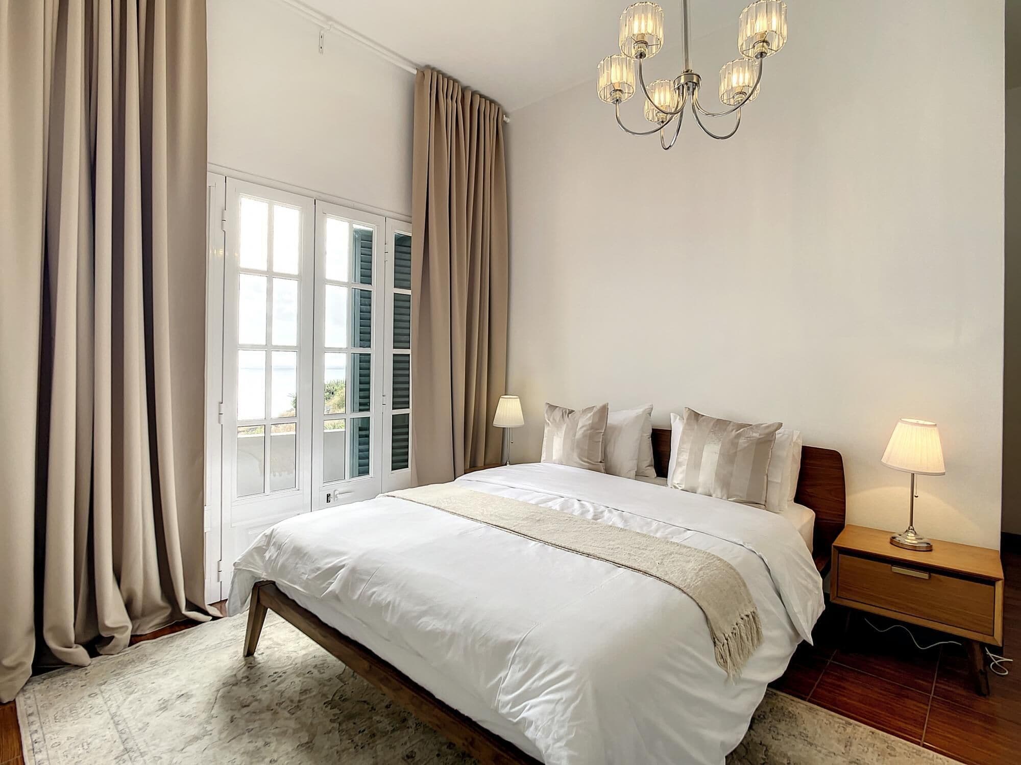 A bedroom with a large bed, two bedside tables with lamps, a chandelier, and a view through a window with curtains.