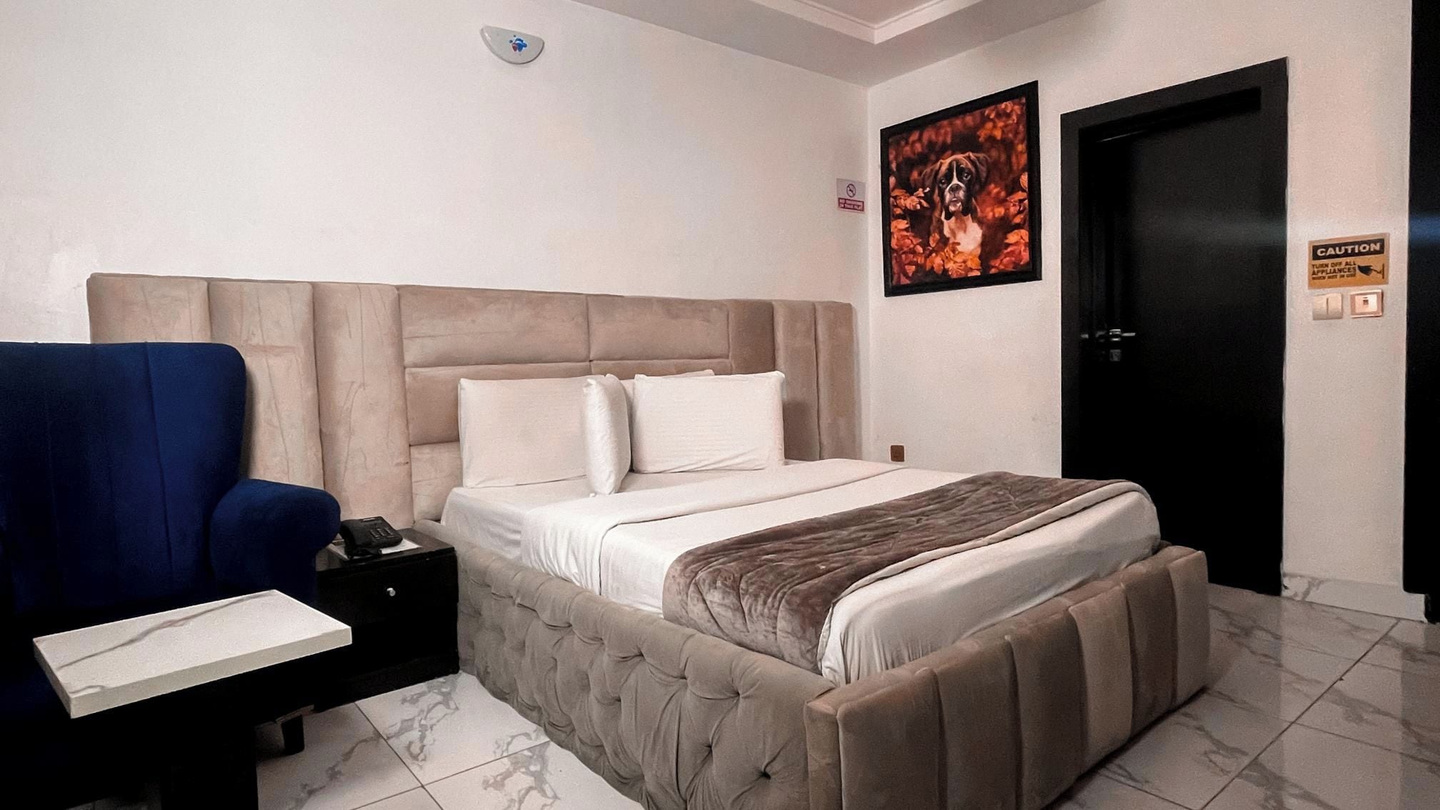 Una camera da letto con un letto, un comodino, una sedia blu, un quadro incorniciato e una porta.