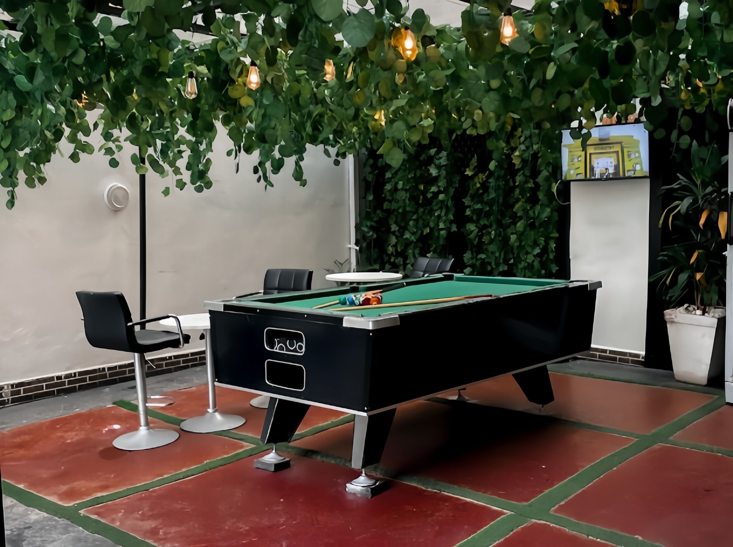 A pool table with a green felt surface, surrounded by a red and green floor. There is a black pool table chair and a white table with a glass top.