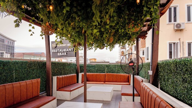 Een terras met zachte banken en tafels, omringd door groen en een bord met restaurant en bar.