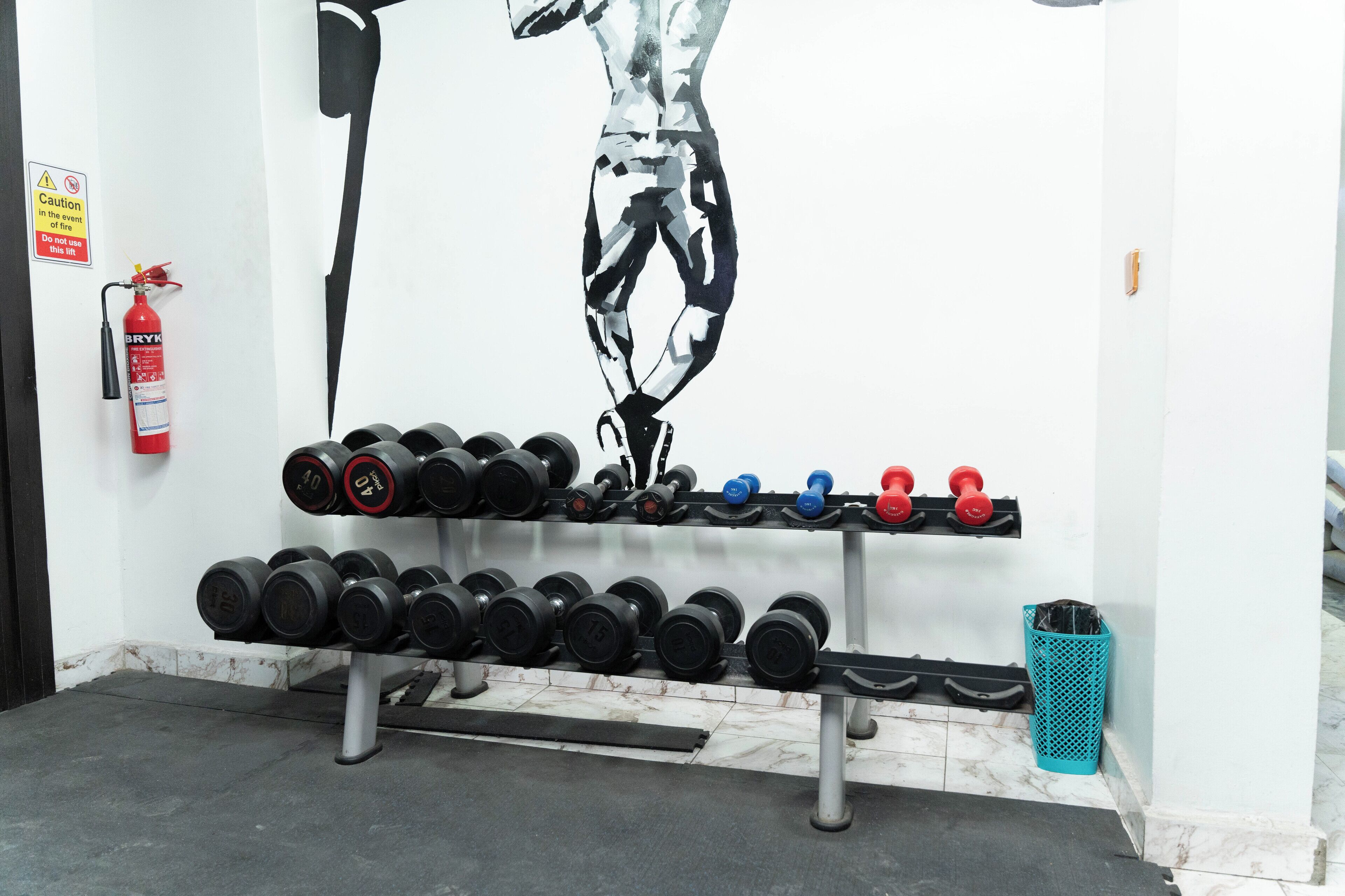 A gym with a wall-mounted fire extinguisher, a wall art of a person jumping, and a rack of dumbbells.