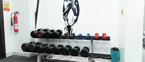 A gym with a wall-mounted fire extinguisher, a wall art of a person jumping, and a rack of dumbbells.