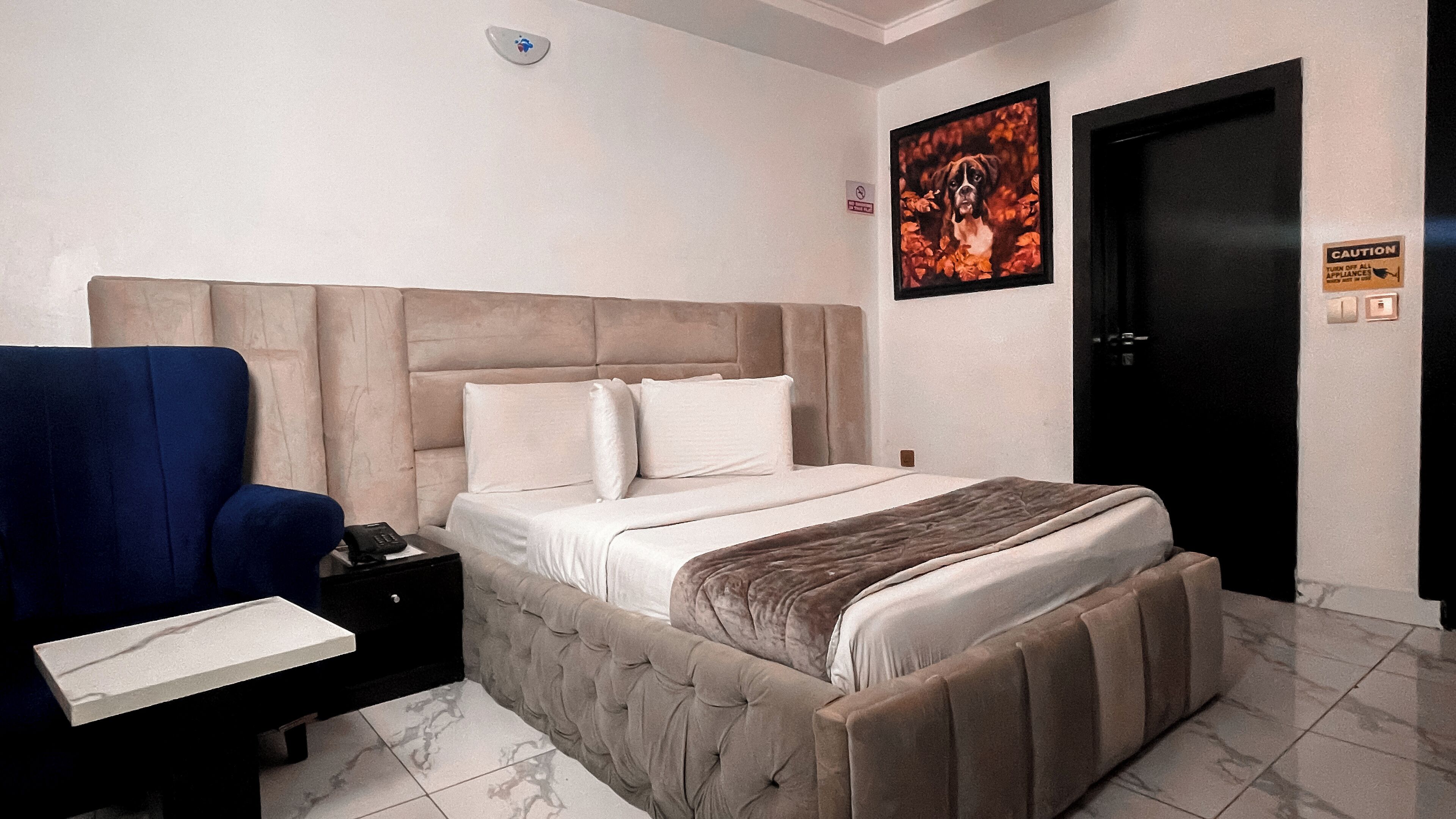 Een slaapkamer met een groot bed, een nachtkastje, een blauwe fauteuil, een ingelijst schilderij aan de muur, en een deur met een waarschuwingsbord.