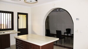 Cozinha com ilha central, armários de madeira escura e bancada branca. Há uma janela com persianas verticais e uma porta com moldura preta.