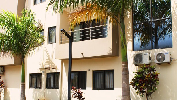 Um edifício com vários andares, unidades de ar condicionado, uma palmeira e uma rua com calçada.
