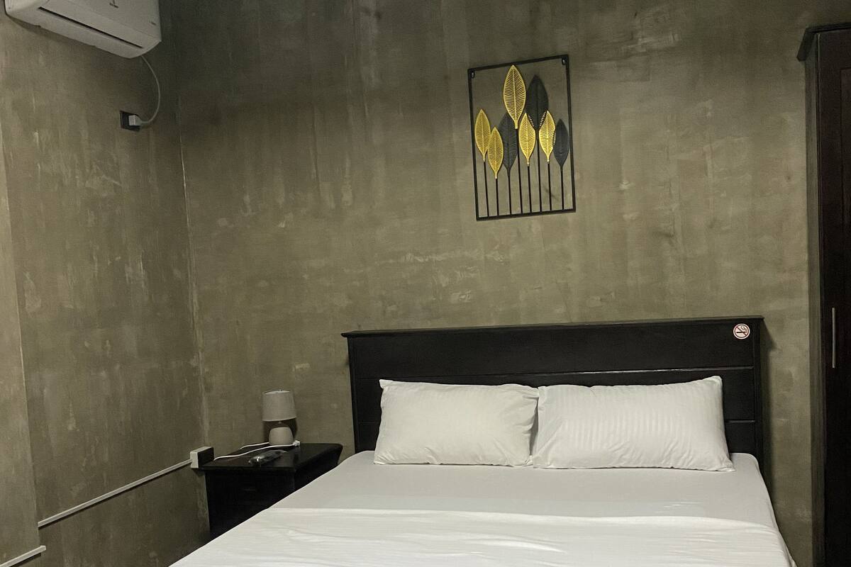 A bedroom with a large bed, a nightstand, a wall-mounted air conditioner, and a decorative wall piece.