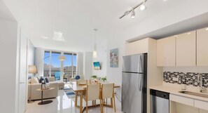 A modern kitchen with a stainless steel refrigerator, a dining area with a table and chairs, and a large window offering a city view.