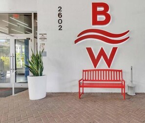 Entrada de un edificio con un banco rojo, un logotipo grande rojo y blanco, y una planta en maceta.