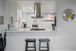 Uma cozinha moderna com armários brancos, coifa de aço inoxidável, micro-ondas e máquina de café. Há dois bancos altos e um espelho decorativo na parede.