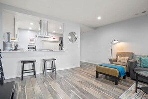 Een moderne keuken met een centraal eiland, twee barkrukken, een zithoek met een bank en fauteuil, en een vloerkleed met een patroon.