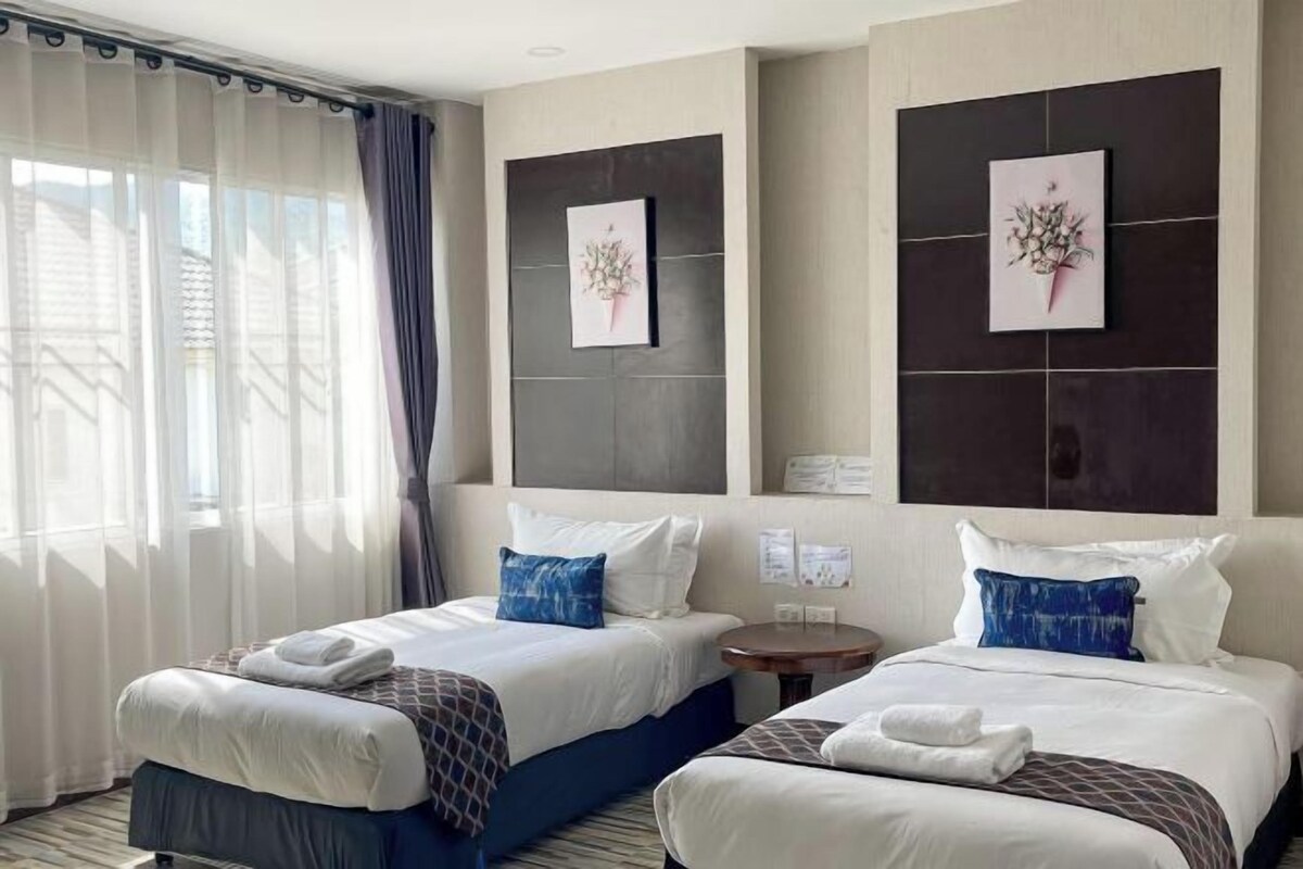 Habitación de hotel con dos camas, una mesita redonda y un ventanal con cortinas traslúcidas.