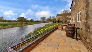 A riverside patio with a brick wall, a wooden bench, a barrel, and a flower bed with yellow flowers. - The Warren (Stamford)