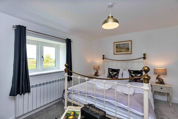 Een slaapkamer met een bed met wit metalen frame, een raam met gordijnen, een radiator, een nachtkastje met lamp en een ingelijst beeld aan de muur.
