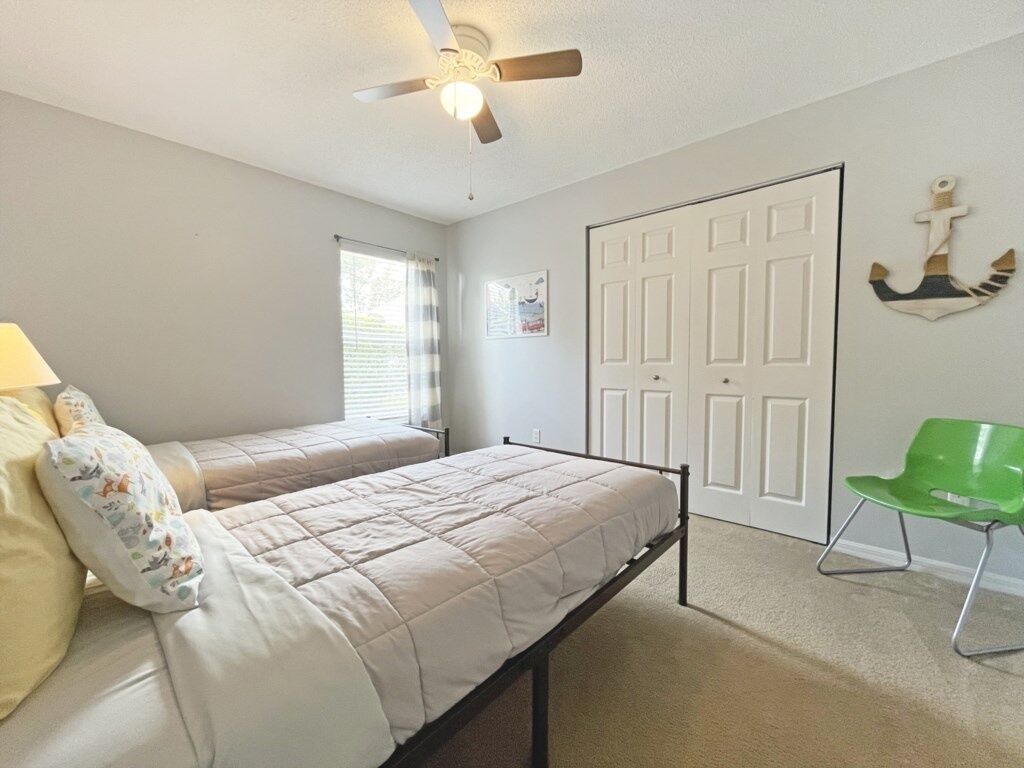 A bedroom with two beds, a ceiling fan, a window with blinds, a door, and a green chair.