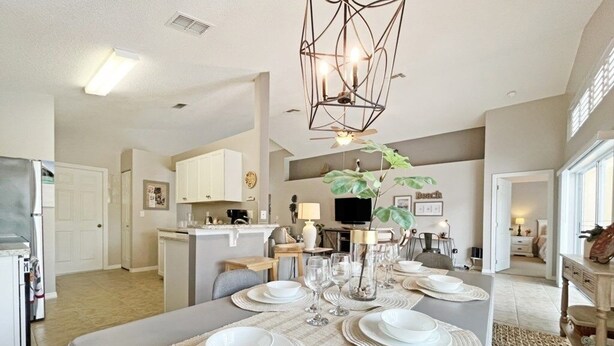 A modern dining area with a rectangular table set for six, surrounded by chairs. There is a kitchen area with white cabinets and a built-in oven to the left. A large plant is placed in the center of the room, and a unique geometric light fixture hangs from the ceiling.