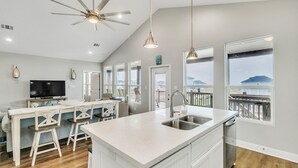 A modern kitchen with a large island, bar stools, a ceiling fan, and large windows offering a view of the outdoors. - Born To Beach 4 Bedroom Home by RedAwning (Port Bolivar)