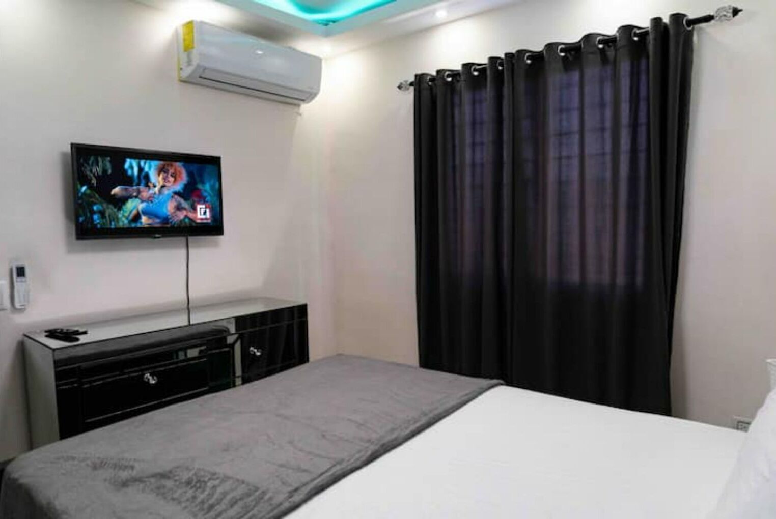 A bedroom with a bed, a TV on a stand, a dresser, and a window with curtains.