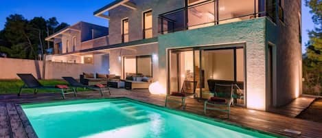 A modern two-story house with a swimming pool, outdoor seating, and large windows.