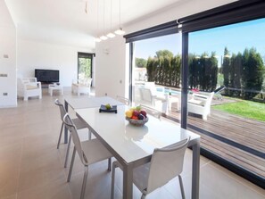 A modern living room with a dining area, a flat-screen TV, and a large window overlooking a pool and greenery. - Villa Luxor by Interhome (L'Ametlla de Mar)