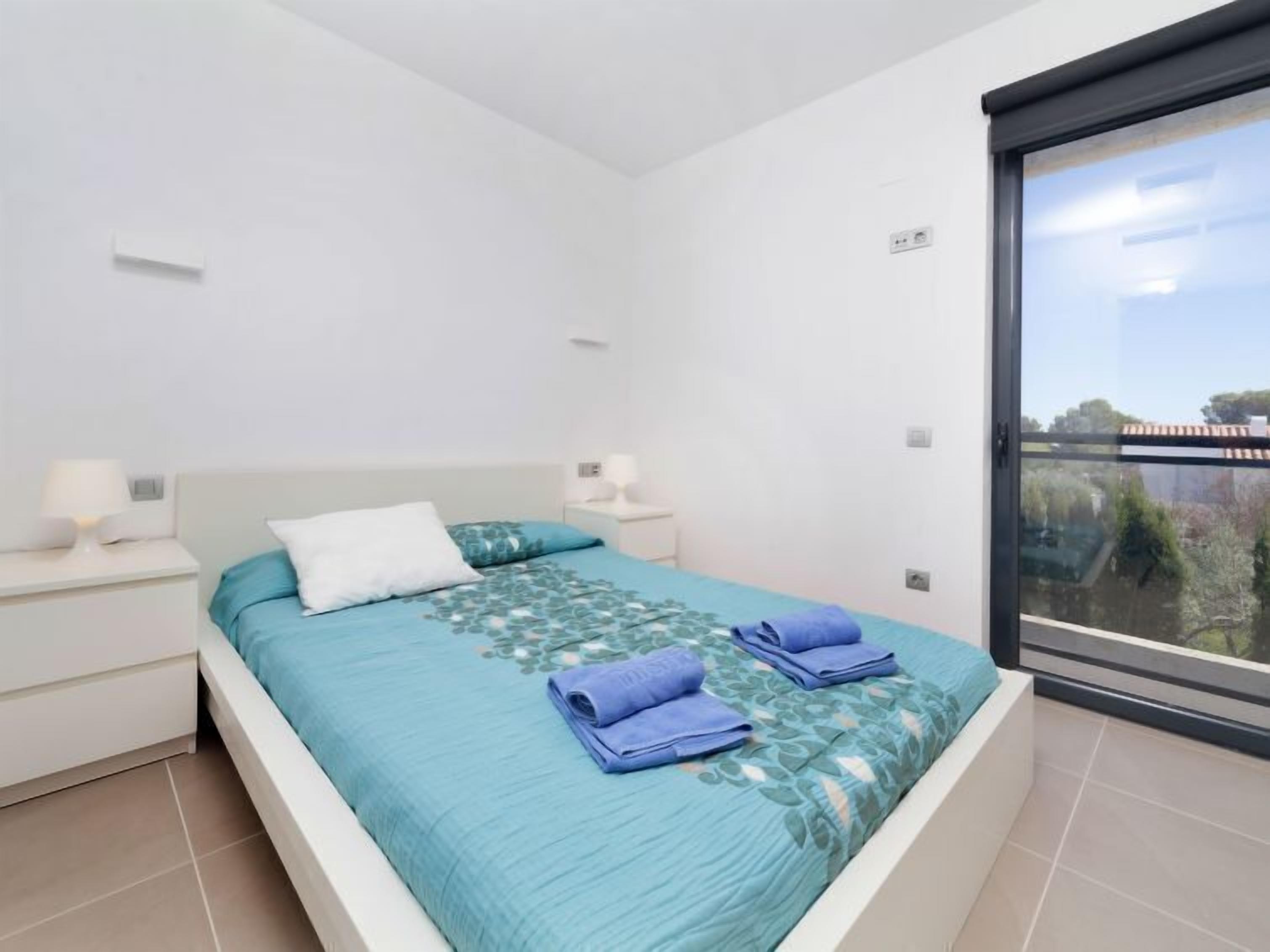 Une chambre moderne avec un lit, des tables de chevet blanches et une grande fenêtre donnant sur un paysage verdoyant et des bâtiments.