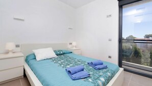 Un dormitorio moderno con cama, mesitas de noche blancas y un amplio ventanal con vistas a zonas verdes y edificios.
