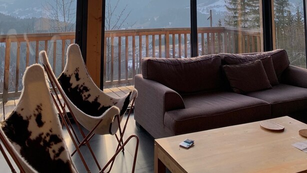 Ein gemütliches Zimmer mit Holzbalkendecke, großen Fenstern mit Bergblick, einem braunen Sofa und einem Tisch mit Stühlen.