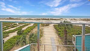 A beach view from a balcony with a clear sky, sandy paths, and vegetation.