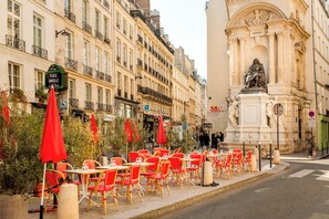 Outdoor seating with tables and chairs, a street with buildings, and a statue.