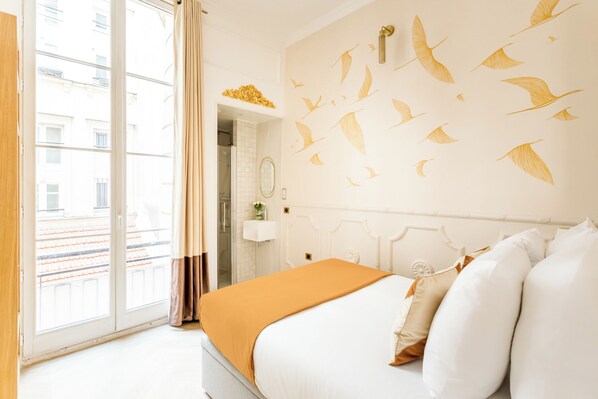 A hotel room with a large window, a bed with white and orange bedding, and a wall with a bird mural.