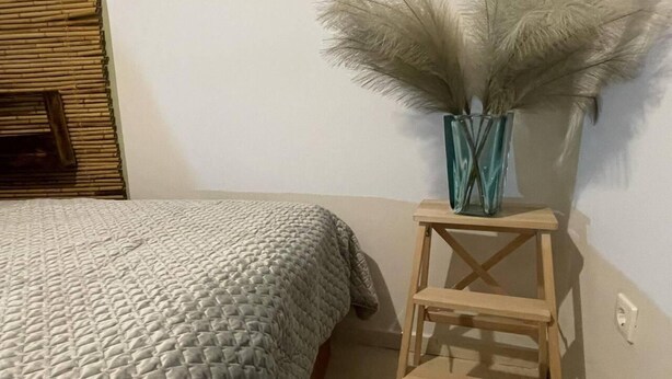 Un dormitorio con una cama, una mesita de noche de madera y un jarrón con plumas de plumero.