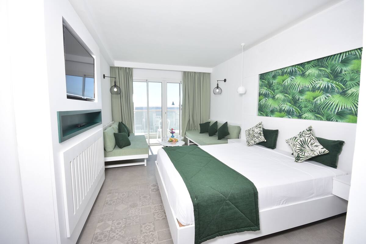 A modern hotel room with a large bed, a sofa, a TV, and a city view.