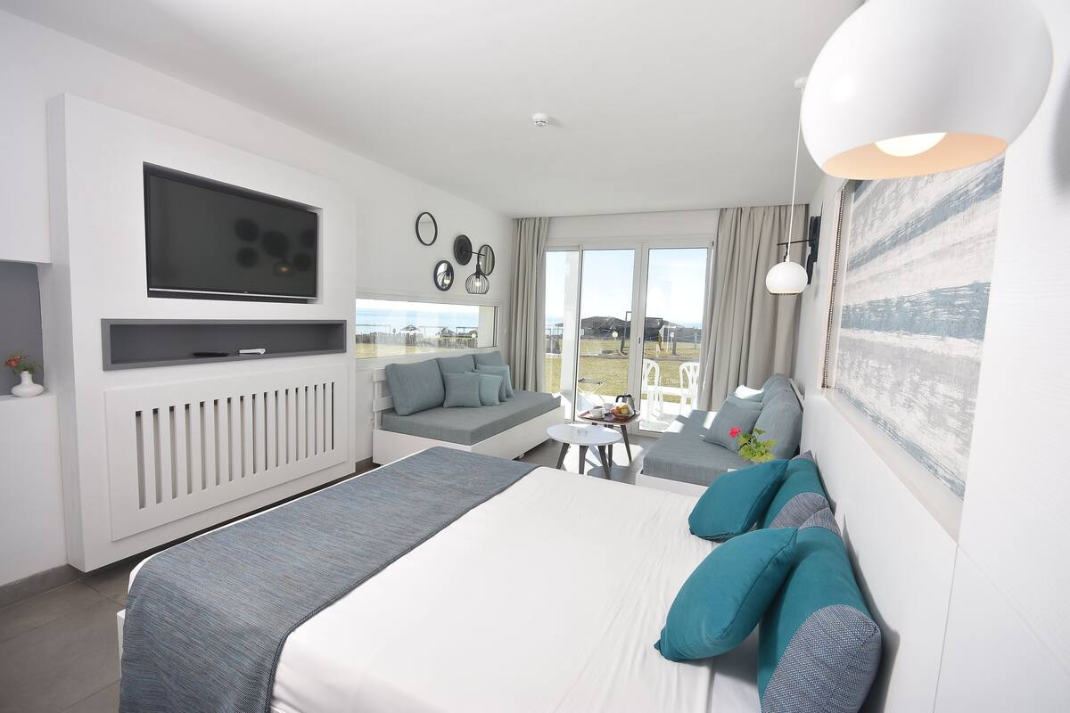 A modern hotel room with a large bed, a sofa, a TV mounted on the wall, and a view of the outdoors.