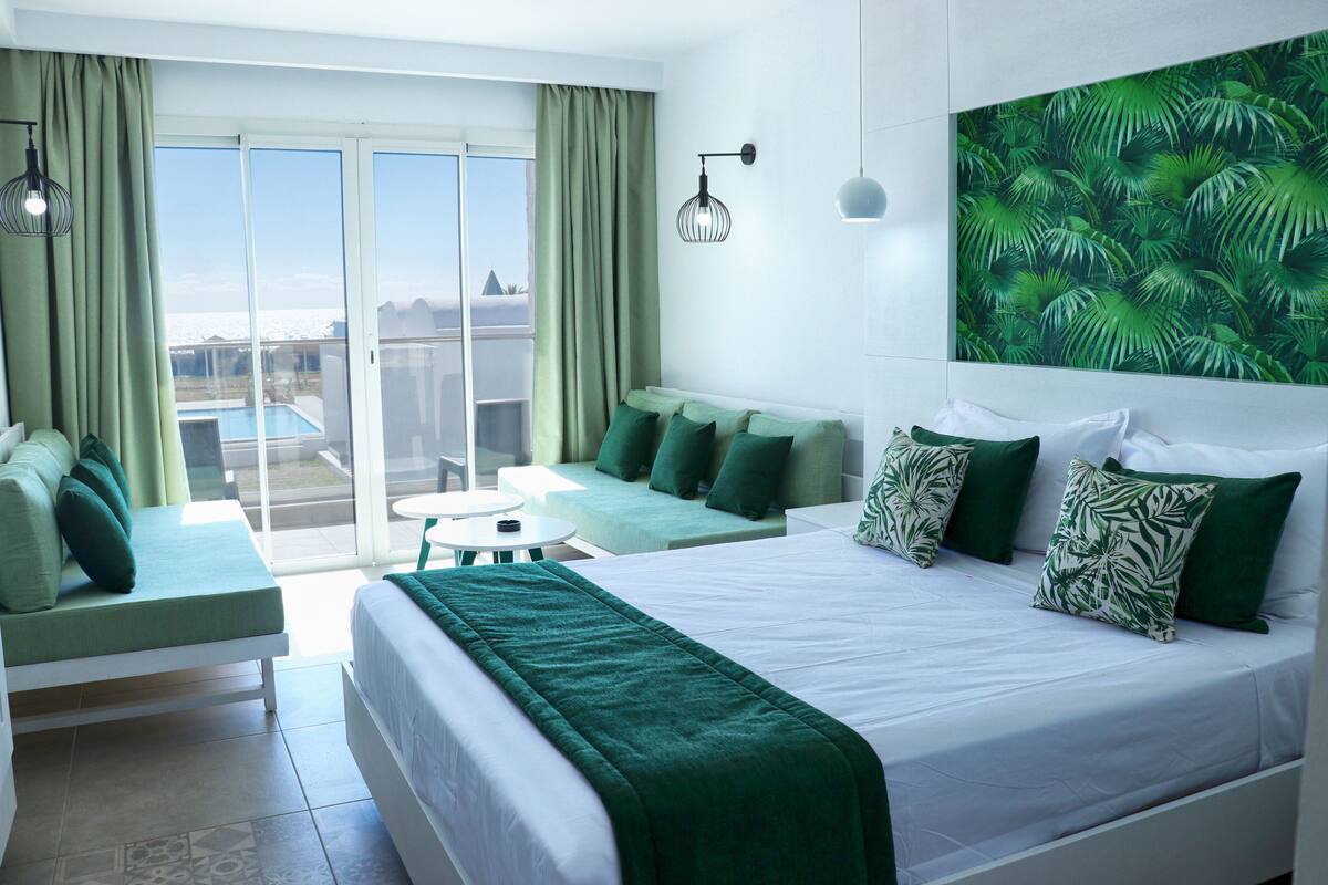 A hotel room with a balcony, a bed with green bedding, a sofa, and a wall-mounted light fixture.