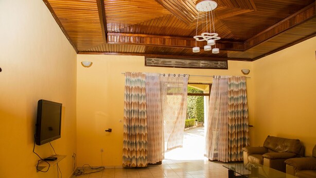 A living room with a wooden ceiling, a flat-screen TV mounted on the wall, a glass coffee table, and a brown leather sofa. - Comfort Apartment  (Kigali)