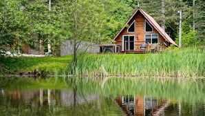 A wooden cabin with a triangular roof, situated by a calm lake surrounded by lush greenery.