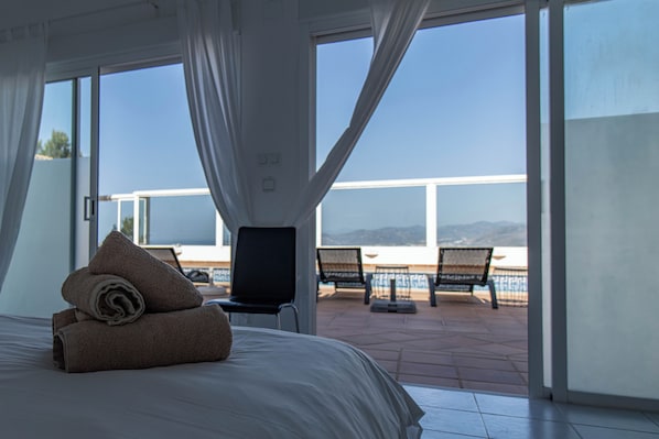 A hotel room with a bed, a TV, and a balcony with chairs and a view of the sea.