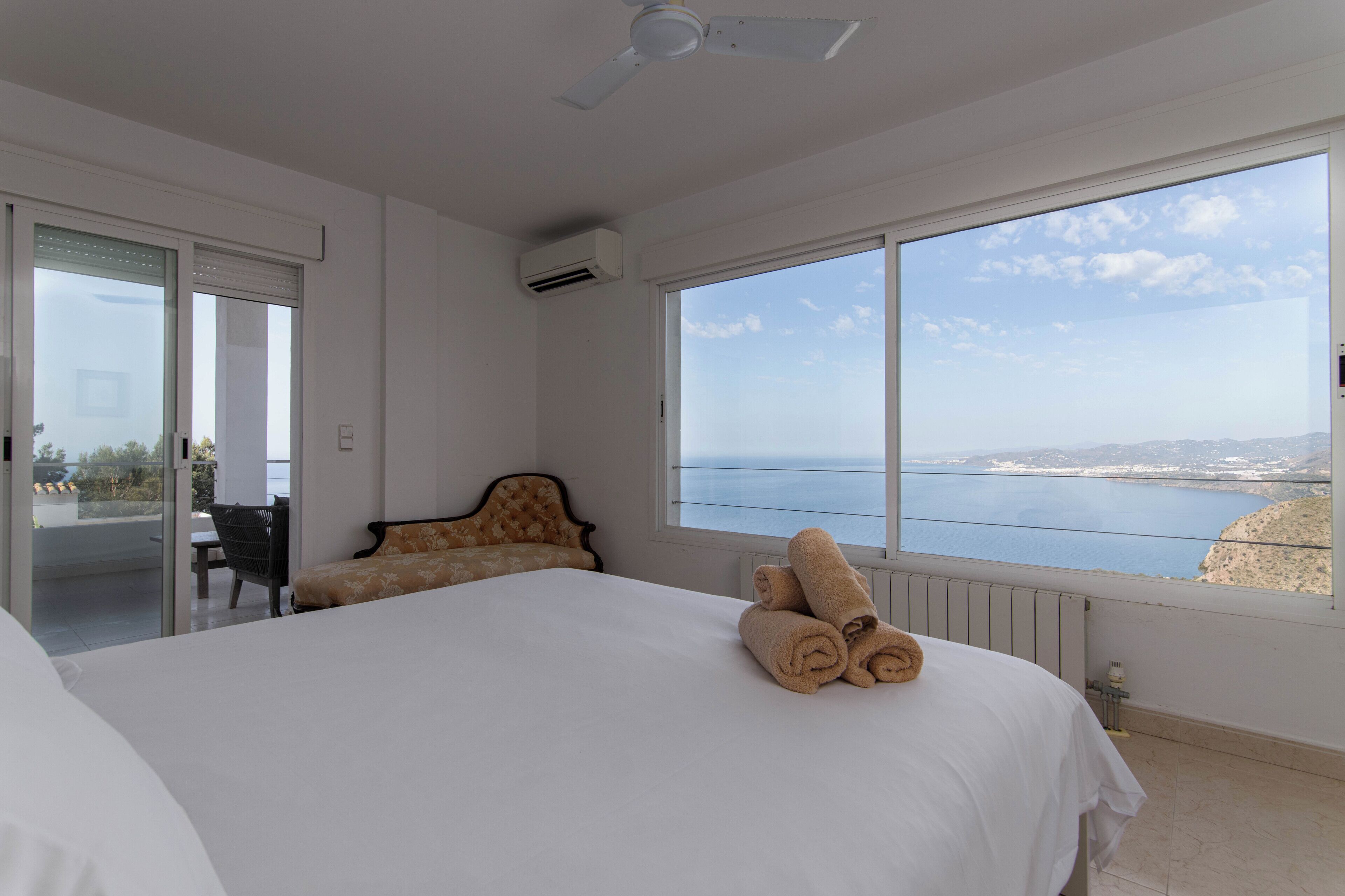 A bedroom with a large bed, a view of the sea, a ceiling fan, and a window with a view of the sea.