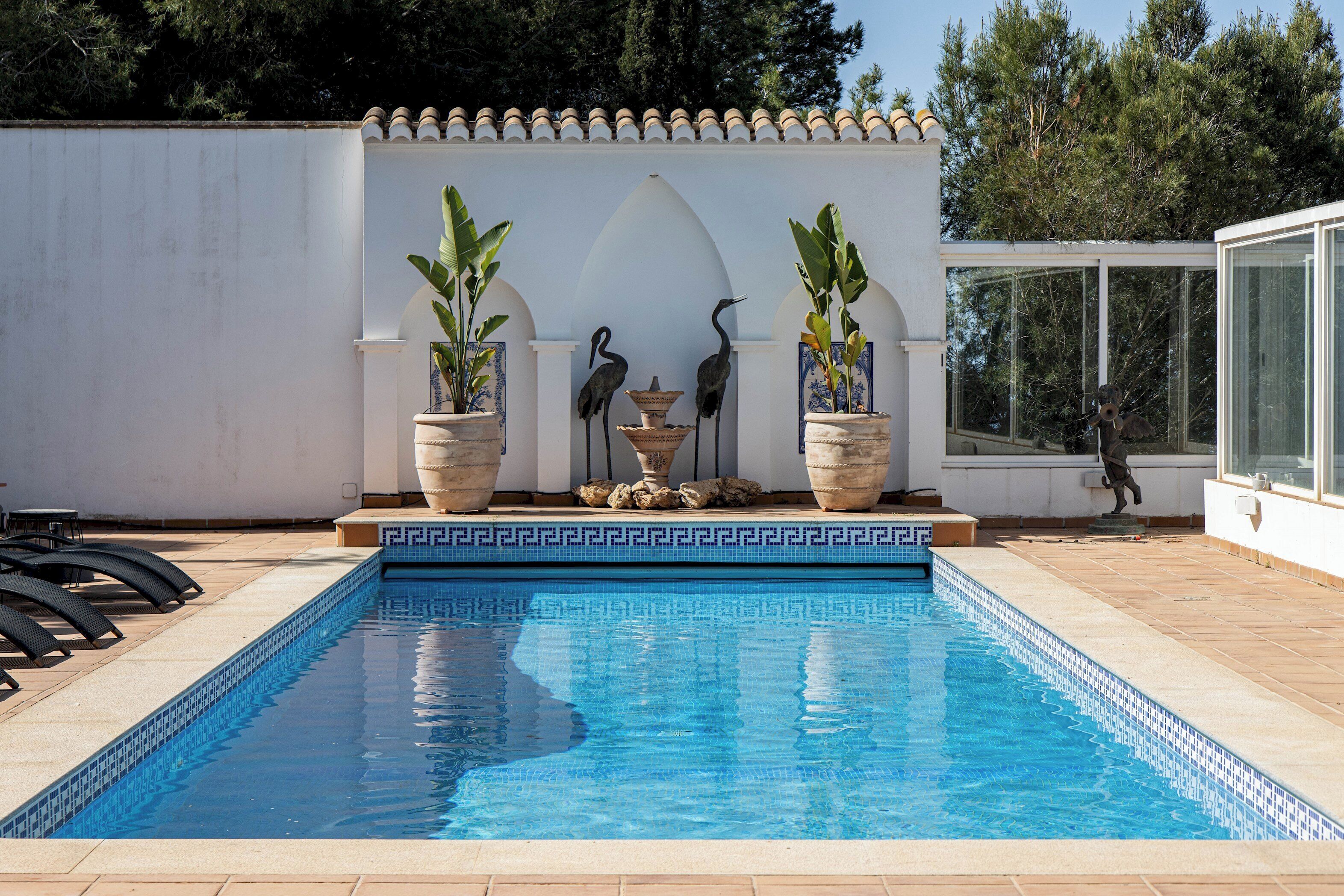 A swimming pool with a tiled edge, surrounded by potted plants and a decorative fountain.