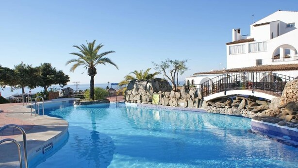 A swimming pool with a stone wall and palm trees, surrounded by a white building with arched windows. - Las Lilas in the heart of Capistrano ,privet  pool (Nerja)