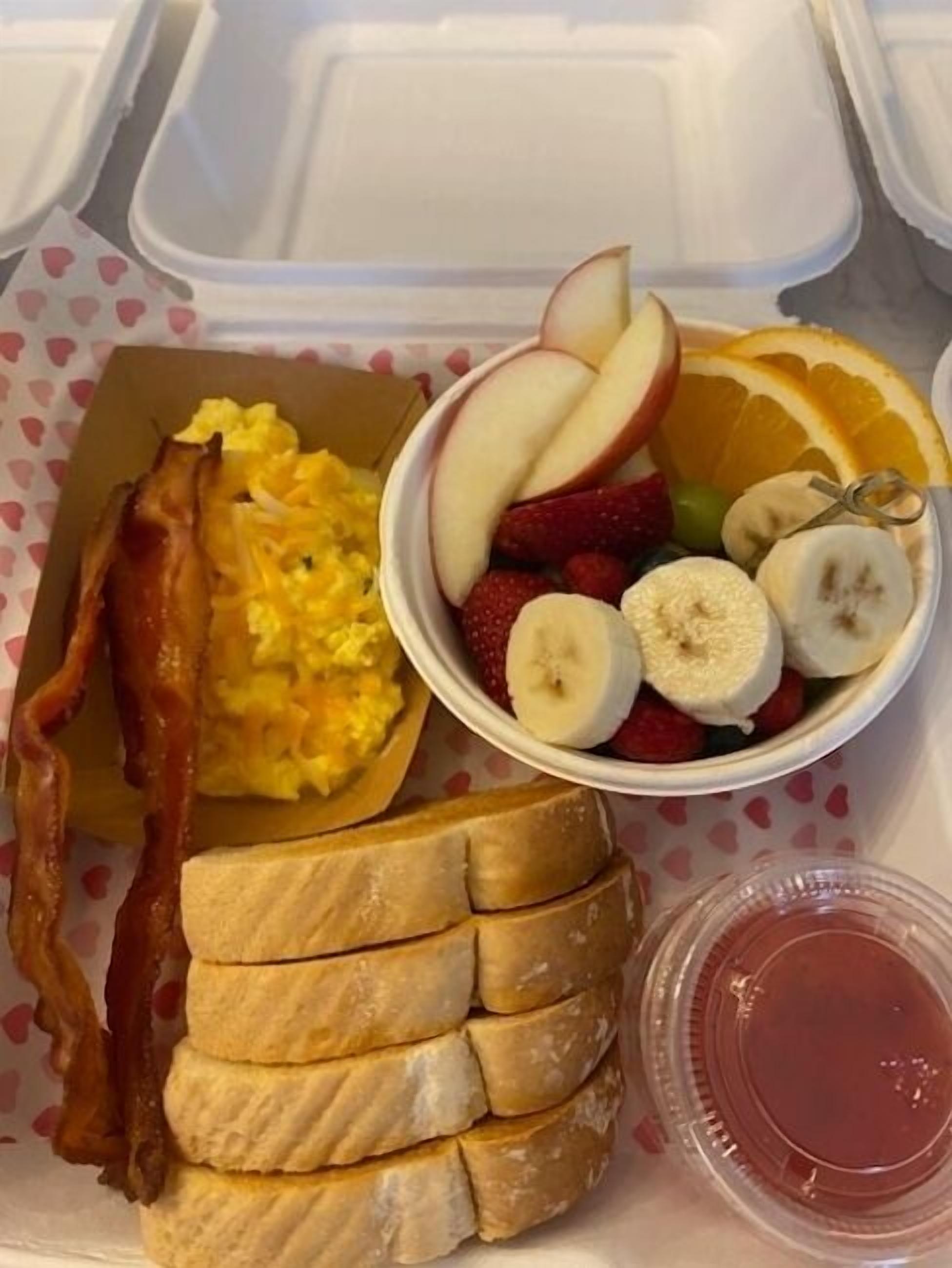 Bandeja de desayuno con huevos revueltos, tocino, tostadas y un bol de fruta con manzanas, fresas, plátanos y naranjas.