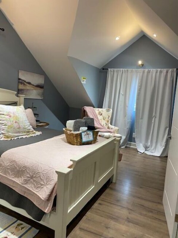 Un dormitorio con techo inclinado, una cama con colcha rosa, una mesita de noche con lámpara, una silla con una cesta encima y una ventana con cortinas.