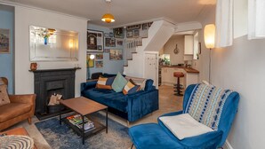 Una sala de estar con chimenea, sofás azules, una mesa de centro y una escalera que lleva a una cocina.