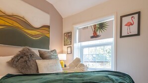 Un dormitorio con cama, almohadas, una lámpara, una ventana con planta y un cuadro de flamenco.
