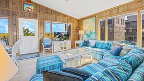 A cozy living room with a striped sofa, a glass coffee table, and a television. - Oceanfront Home with Private Beach Access, Wet Bar, Grill, and Smart TV (Avon)