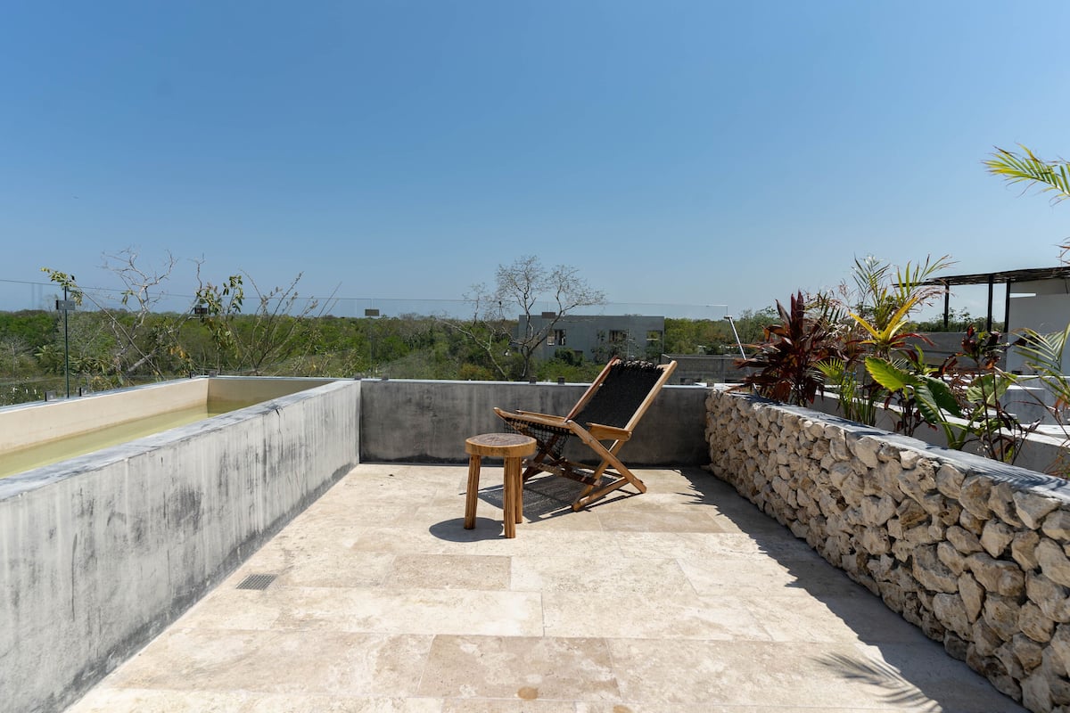 Una terraza en la azotea con una piscina de hormigón, una tumbona de madera, una mesita redonda y un muro de piedra.