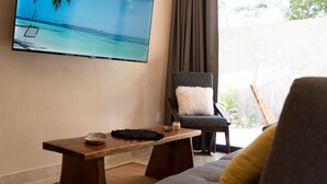 A living room with a flat-screen TV displaying a beach scene, a wooden coffee table, a grey armchair with a white cushion, and a sofa with a yellow cushion. - Xentum Residences - Dreamed Condo (Tulum)