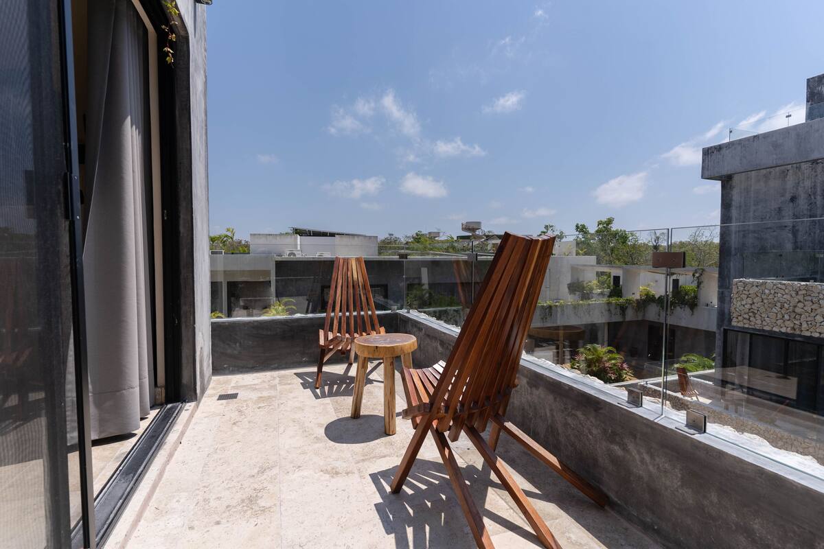 Una terraza en la azotea con barandilla de cristal, dos sillas de madera y una mesita redonda.